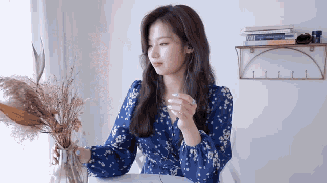 a woman in a blue floral dress is sitting at a table with a vase of dried flowers