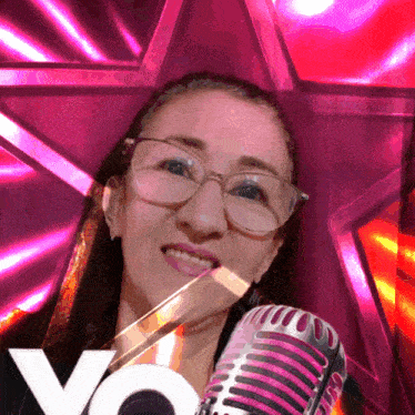 a woman with glasses is holding a microphone in front of a pink background