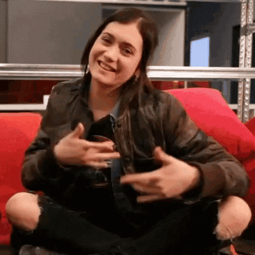 a woman in a leather jacket is sitting on a red couch holding a glass of wine and smiling .