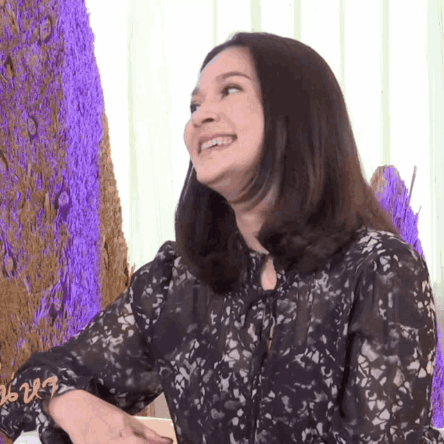 a woman wearing a black lace top smiles in front of purple trees