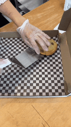 a person wearing gloves is cutting a chocolate chip cookie in a box with a spatula