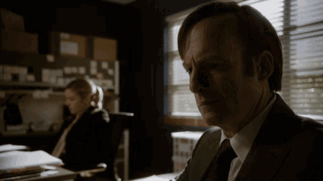 a man in a suit and tie sits in front of a window with blinds