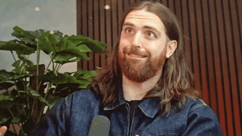 a man with long hair and a beard is talking into a microphone in front of a plant