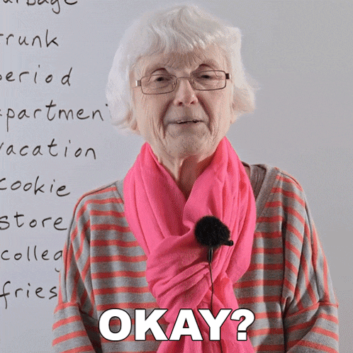 an older woman wearing glasses and a pink scarf says okay in front of a white board