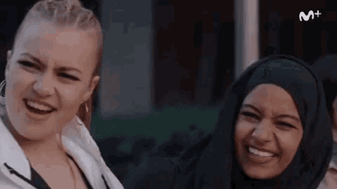 two women are smiling and laughing in front of a sign that says m +