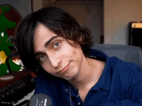 a young man in a blue shirt is looking at the camera with a green christmas tree in the background
