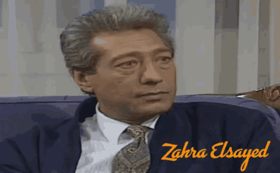 a man in a suit and tie is sitting on a couch with the name zahra elsayed on the bottom right