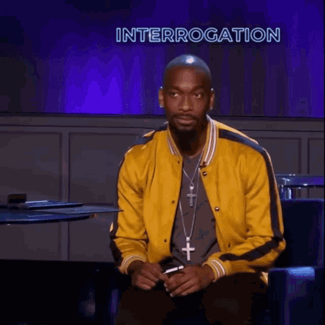 a man wearing a yellow jacket and a cross necklace is sitting in front of a screen that says interrogation