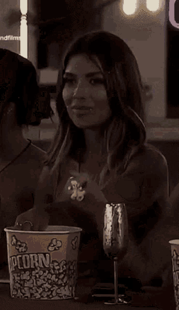 a woman sits at a table with a glass of wine and a bowl of popcorn