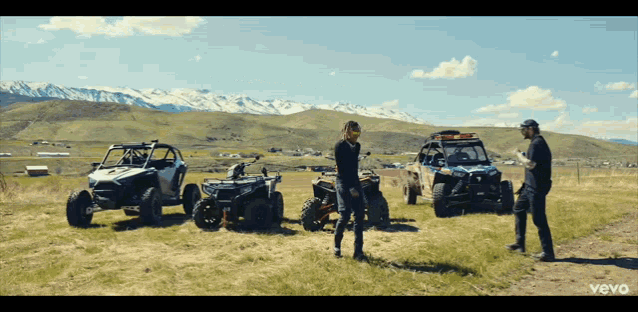 a man in a black jacket stands in a field with four atvs
