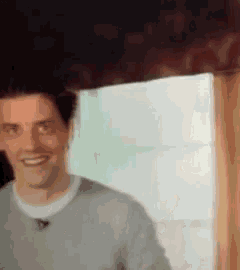 a man in a grey sweater is smiling in front of a window