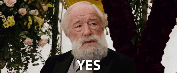 an elderly man with a beard says yes in front of flowers .