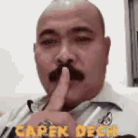 a bald man with a mustache is holding his finger to his mouth while wearing a white shirt .
