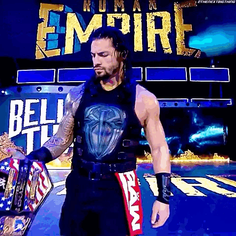 roman reigns is holding a world heavyweight championship belt in front of a sign that says empire .