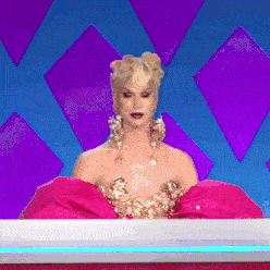 a drag queen is sitting at a table wearing a pink dress and gold earrings .