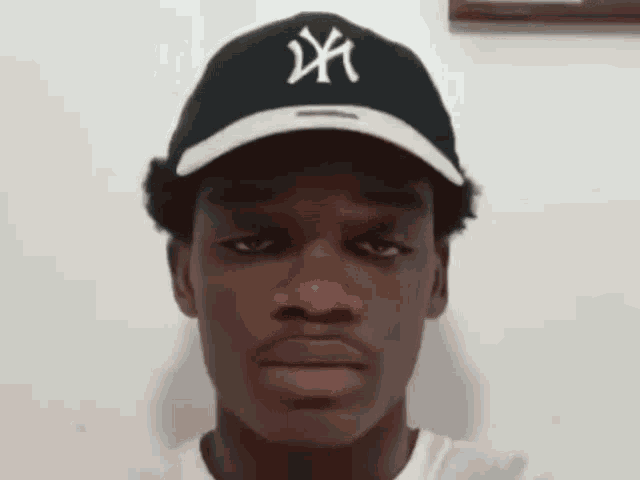 a man wearing a ny hat looks at the camera with a serious look on his face