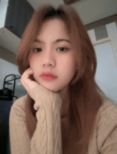 a woman in a tan sweater is taking a selfie with her hand on her chin