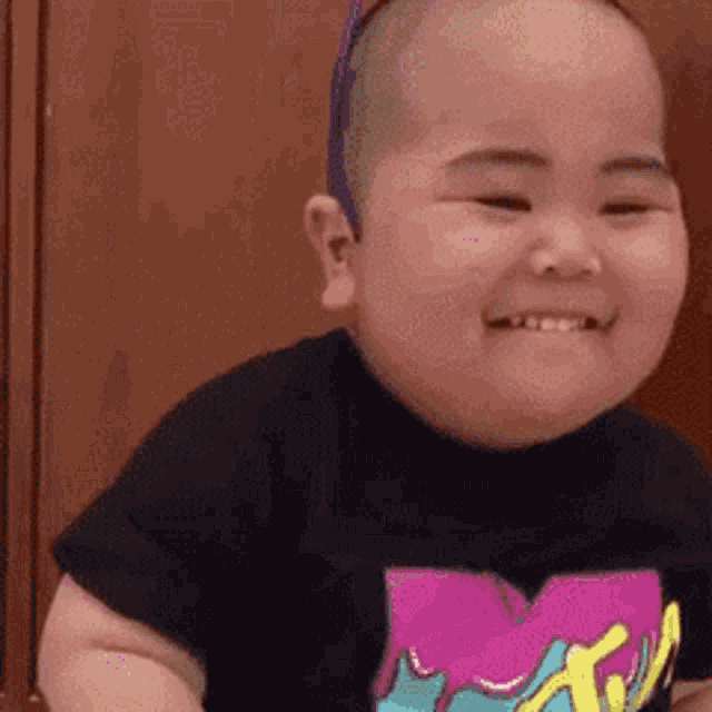 a baby with a shaved head is smiling while wearing a black shirt .