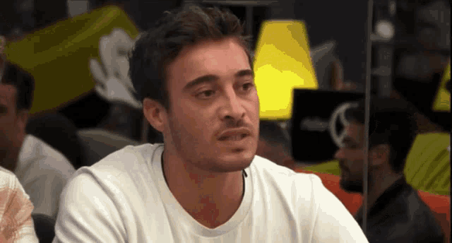 a man wearing a white t-shirt looks at the camera with a serious look on his face