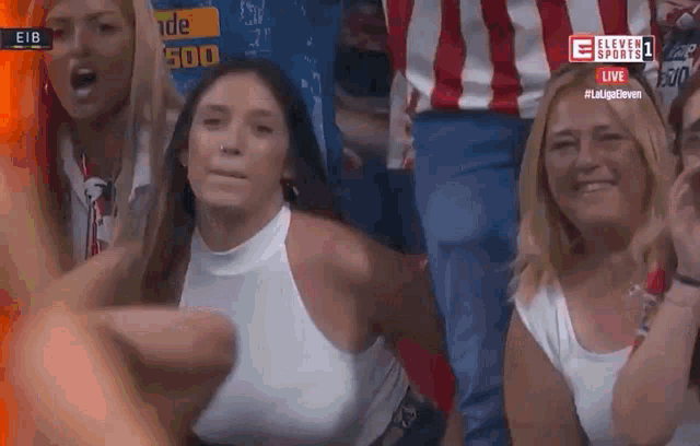 a woman in a white tank top is sitting in a crowd of people watching a soccer game .
