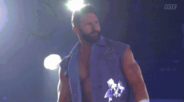 a man with a beard wearing a purple vest is standing in a dark room with the letters fite on the bottom right