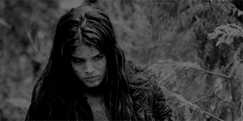 a black and white photo of a woman with long hair standing in a forest .