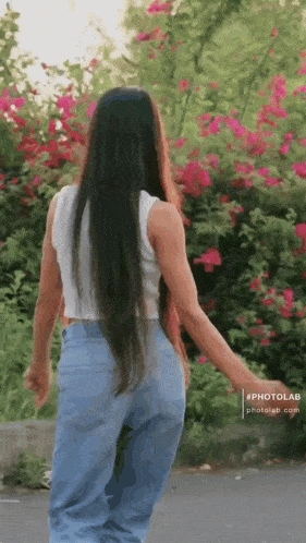 a woman with long hair is walking down a street in front of a bush of pink flowers .