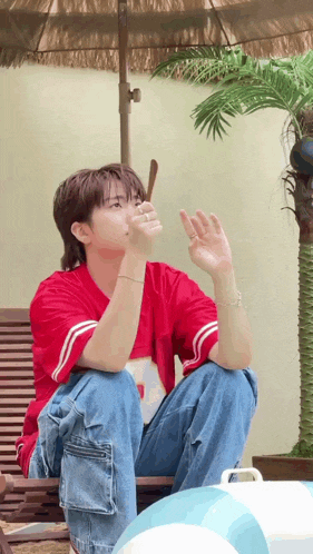 a young man in a red shirt and blue jeans is sitting on a chair under an umbrella