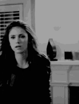 a black and white photo of a woman standing in a living room looking at the camera .