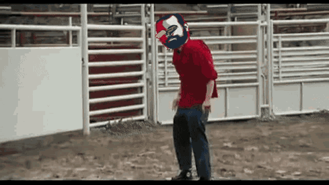 a man in a red shirt is standing in front of a rodeo fence with a mask on his head .