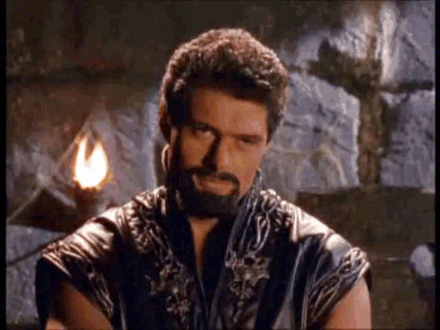 a man with a beard is sitting in a dark room with a torch in the background .