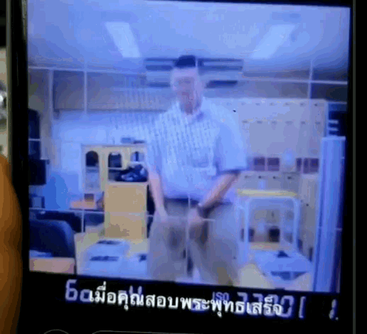 a phone screen shows a man in a plaid shirt standing in an office