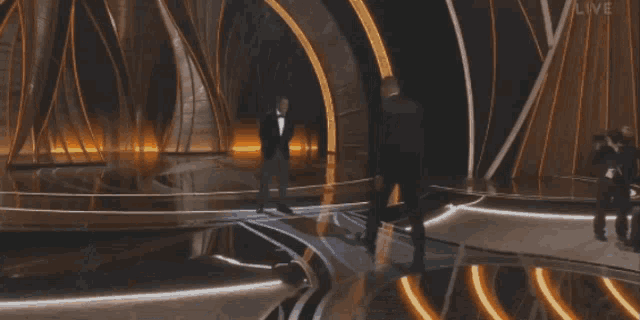 a man in a tuxedo stands on a stage with the word live on the bottom right