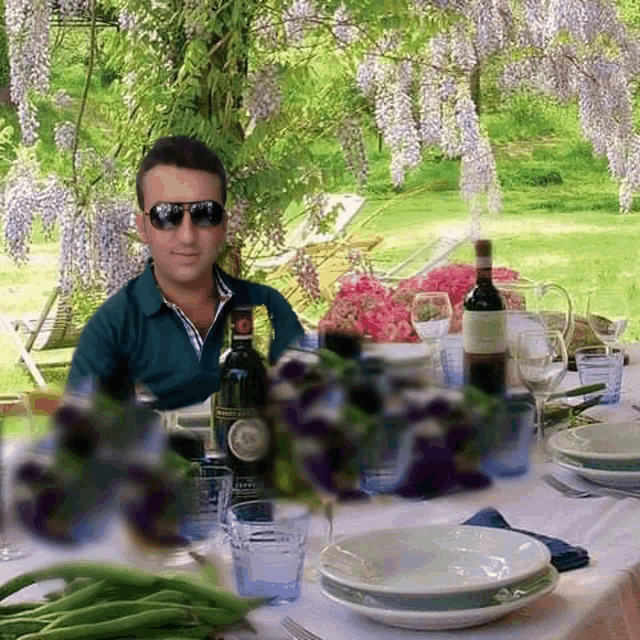 a man wearing sunglasses sits at a table with plates glasses and a bottle of wine with a label on it