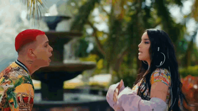 a man with red hair and a woman with long black hair are looking at each other in front of a fountain