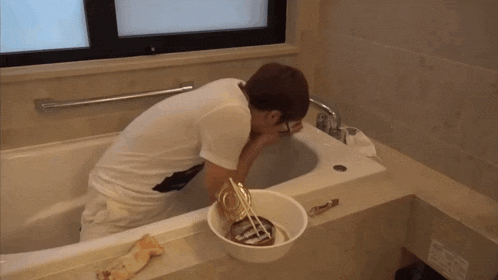 a man washing his face in a bathtub next to a bucket of food