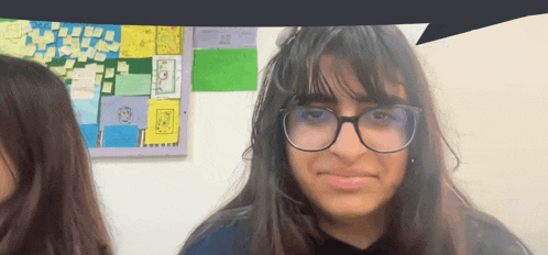 a girl wearing glasses looks at the camera in front of a bulletin board with sticky notes on it