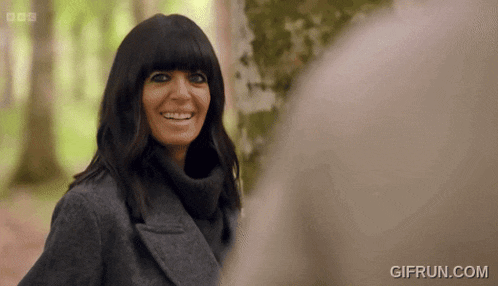 a woman wearing a gray coat and a turtleneck sweater is smiling in a forest .