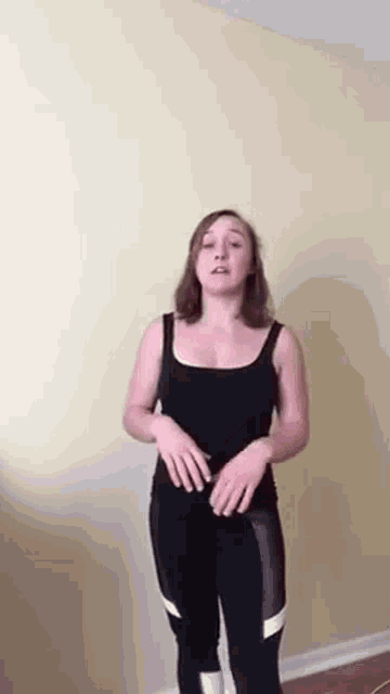 a woman in a black tank top and black leggings is standing in a room with her hands on her hips .
