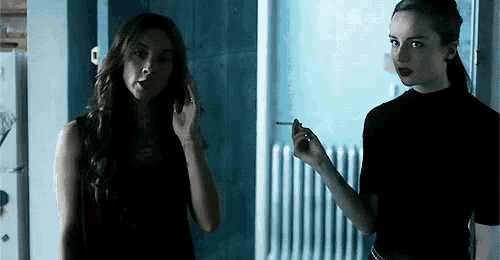 two women are standing next to each other and one is smoking a cigarette while the other is applying makeup .