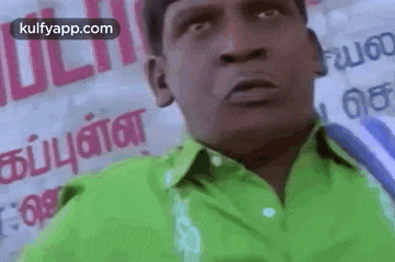 a man in a green shirt is making a funny face while sitting in front of a sign .