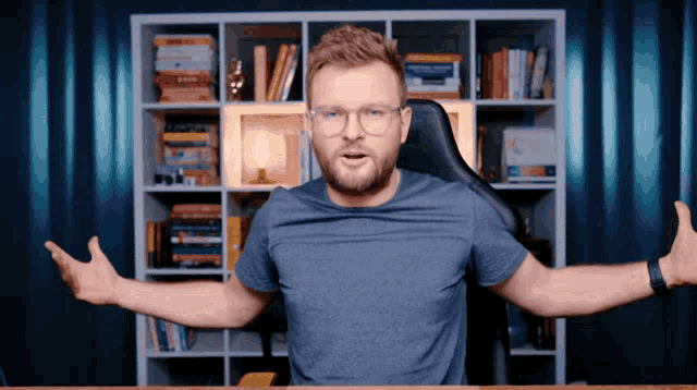 a man wearing glasses and a blue shirt has his arms outstretched in front of a bookshelf
