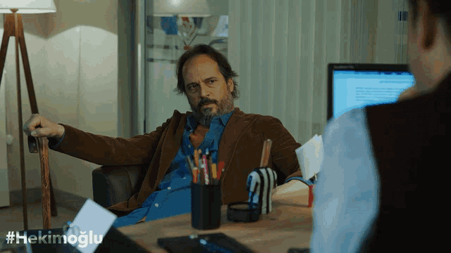 a man with a beard sits at a desk in front of a computer with the hashtag #hekimoglu