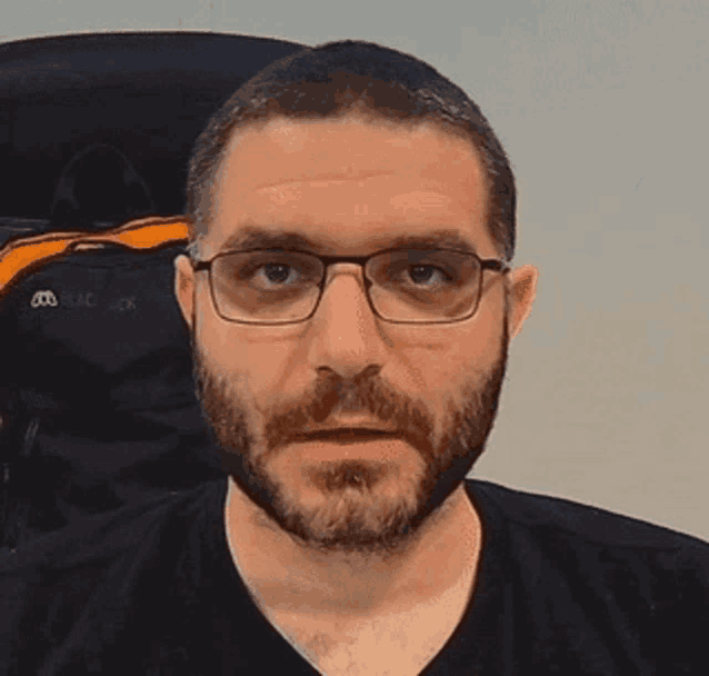 a man with a beard and glasses is wearing a black shirt and looking at the camera .
