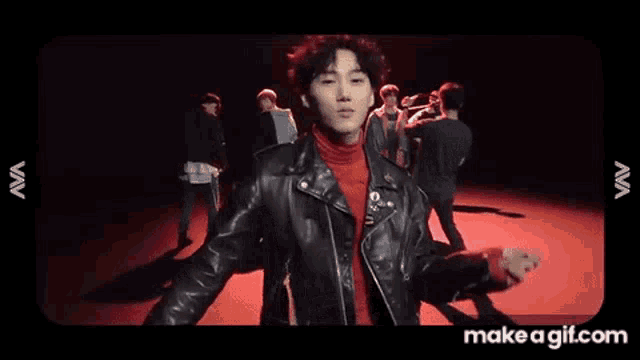 a young man in a black leather jacket and red turtleneck is standing in front of a group of people .