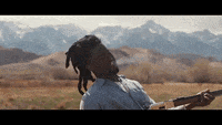 a man is holding a gun in a field with mountains in the background