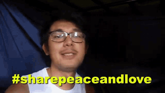 a man wearing glasses and a white tank top is giving a peace sign in front of a sign that says #sharepeaceandlove