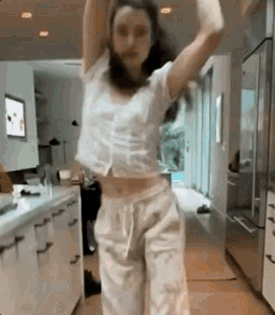 a woman is dancing in a kitchen wearing white pants and a white top .