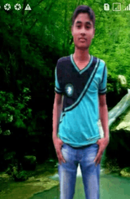 a young man in a blue shirt stands with his hands in his pockets in front of a waterfall