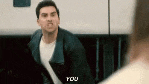 a man in a leather jacket is standing in front of a white wall and yelling at someone .
